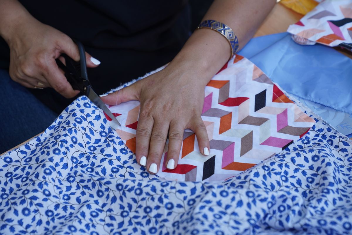 Hands using black scissors to cut blue floral fabric beside a multicoloured geometric fabric piece