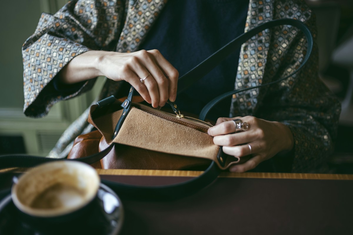 Hands opening the zip of a brown handbag at a café table beside a cup of coffee