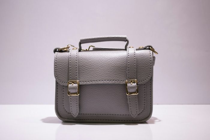 A grey satchel with a top handle and two gold-coloured buckle details on a pale surface