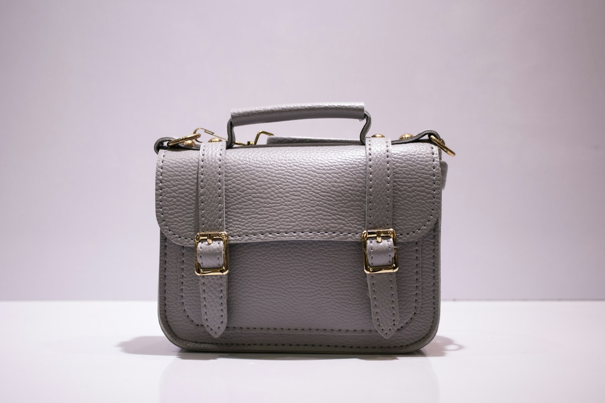 A grey satchel with a top handle and two gold-coloured buckle details on a pale surface