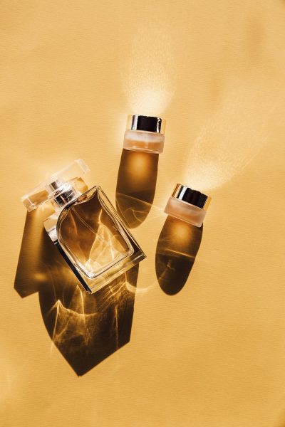 Glass perfume bottle and two small cosmetic jars casting shadows on a yellow surface