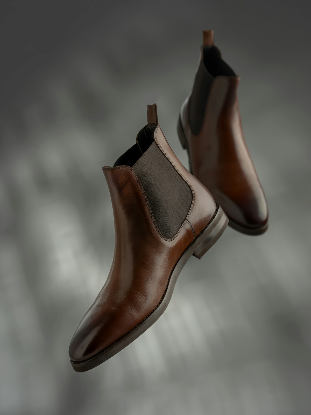 Polished brown Chelsea boots with elastic side panels against a grey background