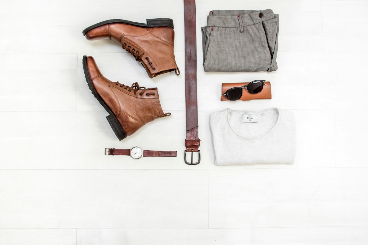 Brown lace-up boots arranged beside grey trousers, a white jumper, belt, watch and sunglasses